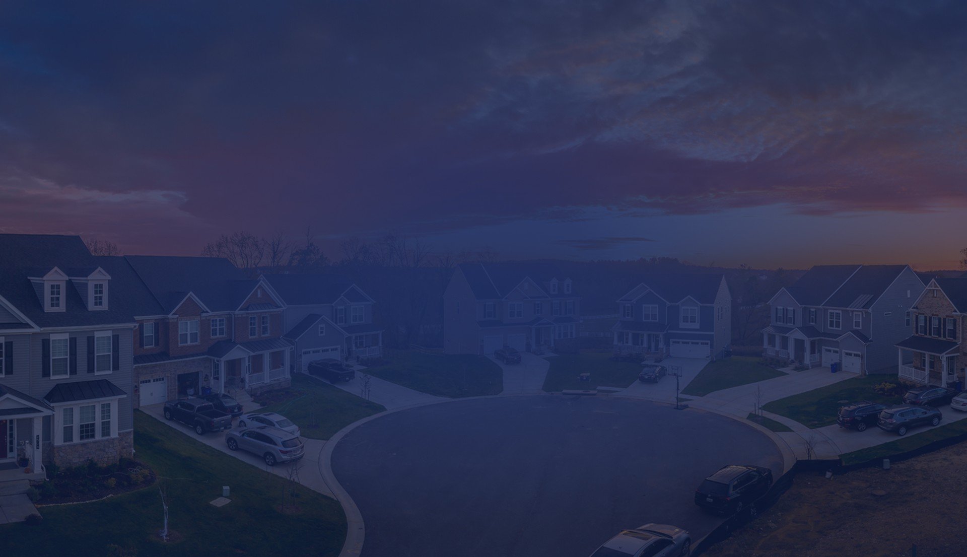 An aerial view of a suburban cul-de-sac lined with new homes under a vivid orange and pink sunset sky.